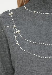 Pulover gri din tricot cu guler înalt, decorat cu multiple șiruri de perle false albe, mari și mici, dispuse în modele drapate.