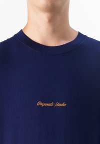 Mornarsko plava košulja s izvezenim logotipom "Originals Studio" u zlatnoj boji na prsima, usko okvirena oko izreza.