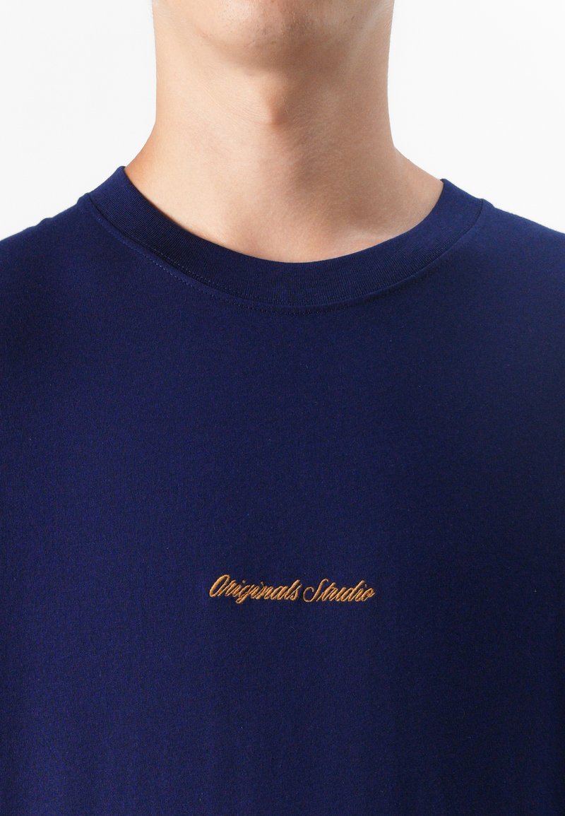Granatowa koszula z wyszywanym złotym logo "Originals Studio" na klatce piersiowej, blisko obramowana wokół dekoltu.