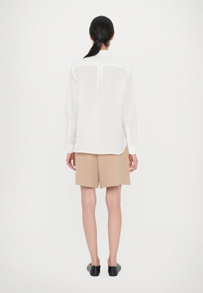 White long-sleeve shirt with a back yoke detail, paired with beige tailored shorts and black slip-on shoes, shown from the back.