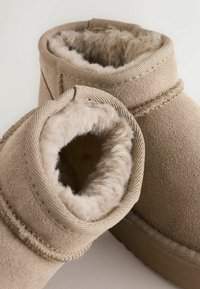 Beżowe zamszowe botki na kostkę z puszystym futerkiem wewnątrz, zaokrąglonym kołnierzem i grubą gumową podeszwą. Posiadają widoczne szwy oraz teksturowane akcenty.