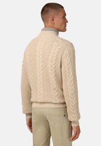 Beige kabelstickad tröja med ribbad krage och muddar. Har ett upphöjt mönster längs ryggen och sidorna, vilket visar på textilens struktur.