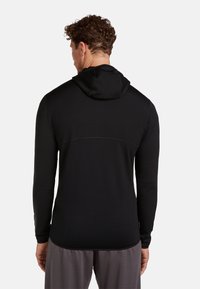 Uomo con capelli ricci che indossa una felpa con cappuccio nera a maniche lunghe e pantaloni grigi, mostrato di spalle su sfondo bianco.