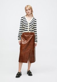 Gestreifter Cardigan in Creme und Marineblau mit Knöpfen, kombiniert mit einem braunen Lederwickelrock. Schwarze Loafer runden das Outfit ab.