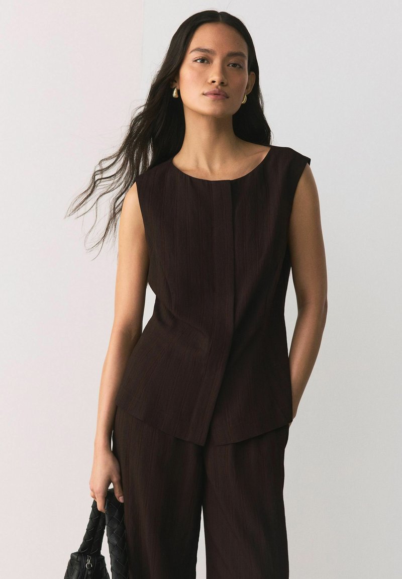 Donna con lunghi capelli scuri che indossa un top smanicato marrone scuro e pantaloni abbinati, mentre tiene una borsa intrecciata nera su uno sfondo chiaro e uniforme.