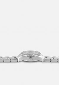 Montre en métal argenté avec un boîtier rond, une lunette texturée et un bracelet à maillons. Cadran blanc avec huit marqueurs et une couronne sur le côté.