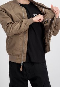 Giubbotto bomber color khaki con un esterno liscio, polsini a coste e chiusura con zip. La maglietta nera sottostante presenta un logo.