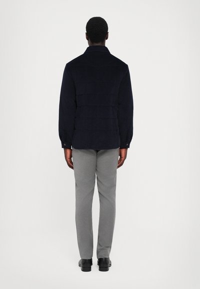 Dunkelblaue Cordjacke mit strukturiertem Design und geknöpften Manschetten, kombiniert mit hellgrauen Hosen und schwarzen Schuhen. Rückansicht.