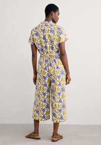 Blommig jumpsuit med korta ärmar och bälte i midjan, med gula, lila och gröna mönster på en ljus bakgrund. Korta ben.