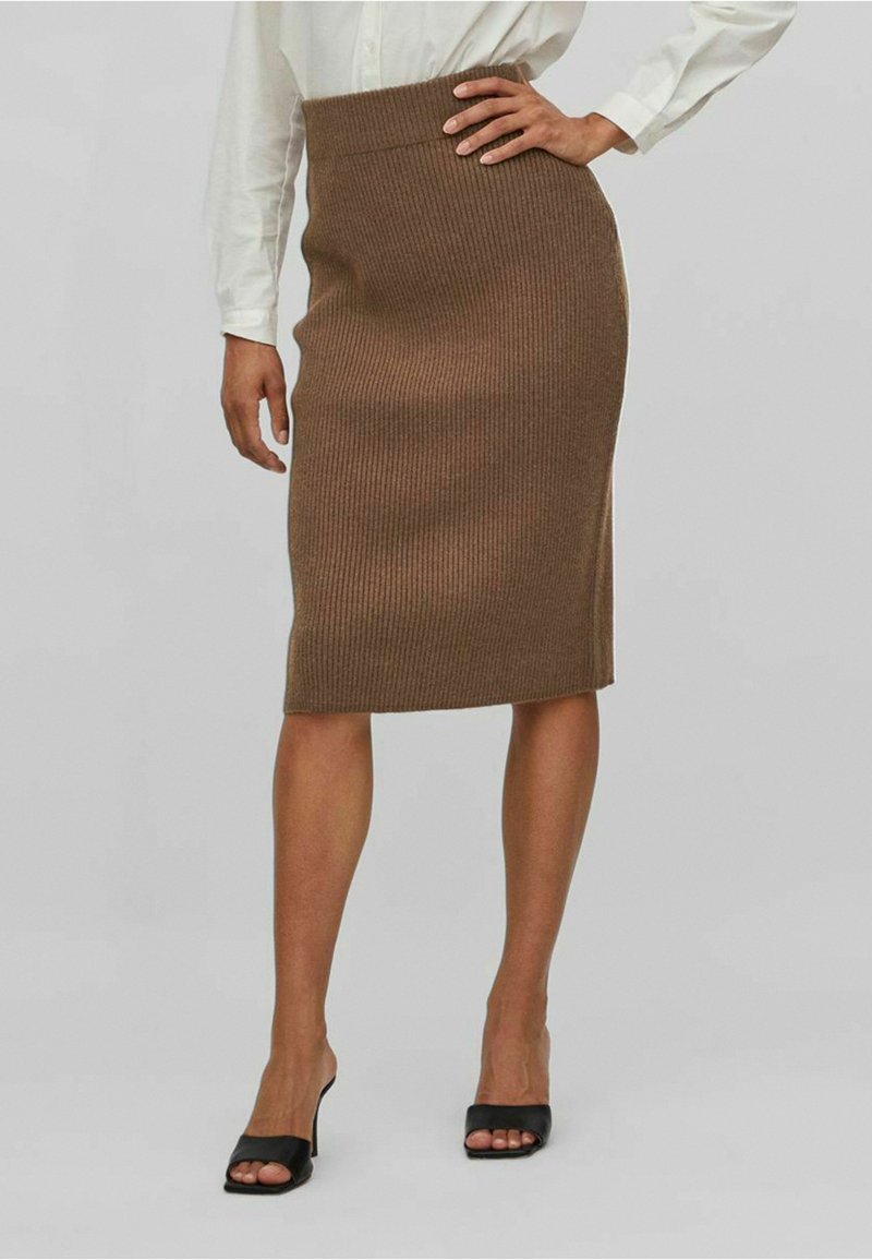 Femme portant une jupe crayon marron en maille côtelée, une chemise blanche à manches longues et des talons ouverts noirs, posant avec une main sur la hanche.