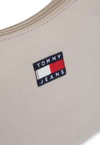 Μπεζ υφασμάτινη τσάντα με υφή επιφάνειας, διακοσμημένη με μπλε και κόκκινο κεντημένο λογότυπο Tommy Jeans. Κλείνει με φερμουάρ.