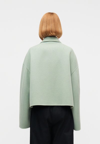 Γυναίκα με ίσια, ξανθά μαλλιά μήκους μέχρι τους ώμους, που φοράει ένα άνετο, ανοιχτοπράσινο μπουφάν και μαύρο παντελόνι, σε θέση που γυρίζει την πλάτη της, πάνω σε λευκό φόντο.