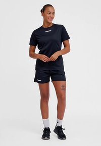 Czarny sportowy strój składający się z koszulki z krótkim rękawem i spodenek. Materiał jest gładki, z logo na klatce piersiowej i udzie. Akcesoria to białe skarpetki i czarne trampki.