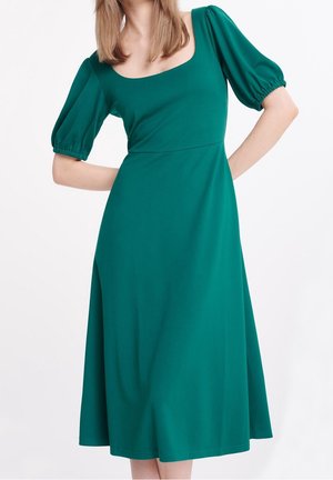 Vrouw in een teal midi-jurk met opgeblazen mouwen tot aan de ellebogen en een vierkante halslijn, staand met handen in de zij.