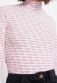 Maglietta a maniche lunghe con colletto a dolcevita di colore bianco, con ripetuto testo rosso "Calvin Klein". Il tessuto ha una consistenza liscia e una silhouette aderente.