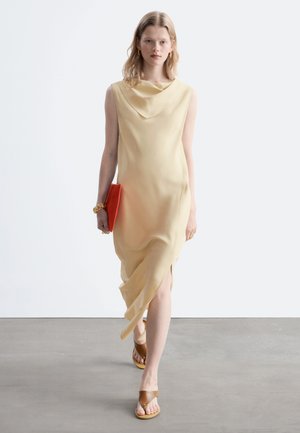 Femme marchant, portant une robe beige sans manches, tenant une pochette rouge, avec des sandales marron sur un sol gris et un fond blanc.