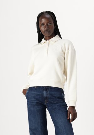 Crème kleur sweatshirt met een polo kraag en geborduurd logo, gecombineerd met donkerblauwe spijkerbroek met rechte snit en zakken.