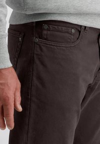 Pantalon marron foncé avec une texture lisse, doté d'accents métalliques sur les poches et les coutures. Un pull gris est visible en haut.
