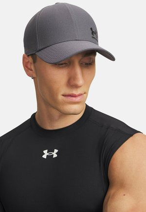 Young man wearing a gray cap and black sleeveless Under Armour shirt looking down and to the side against a white background.