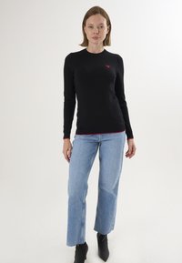 Haut noir à manches longues avec un col rond et un détail en forme de cœur rouge. Associé à un jean flare bleu clair et des bottines noires.