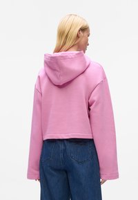 Gekürzter pinker Hoodie mit Kapuze, langen Ärmeln und glatter Textur; kombiniert mit dunkelblauen Jeans aus Denim mit Gesäßtaschen.