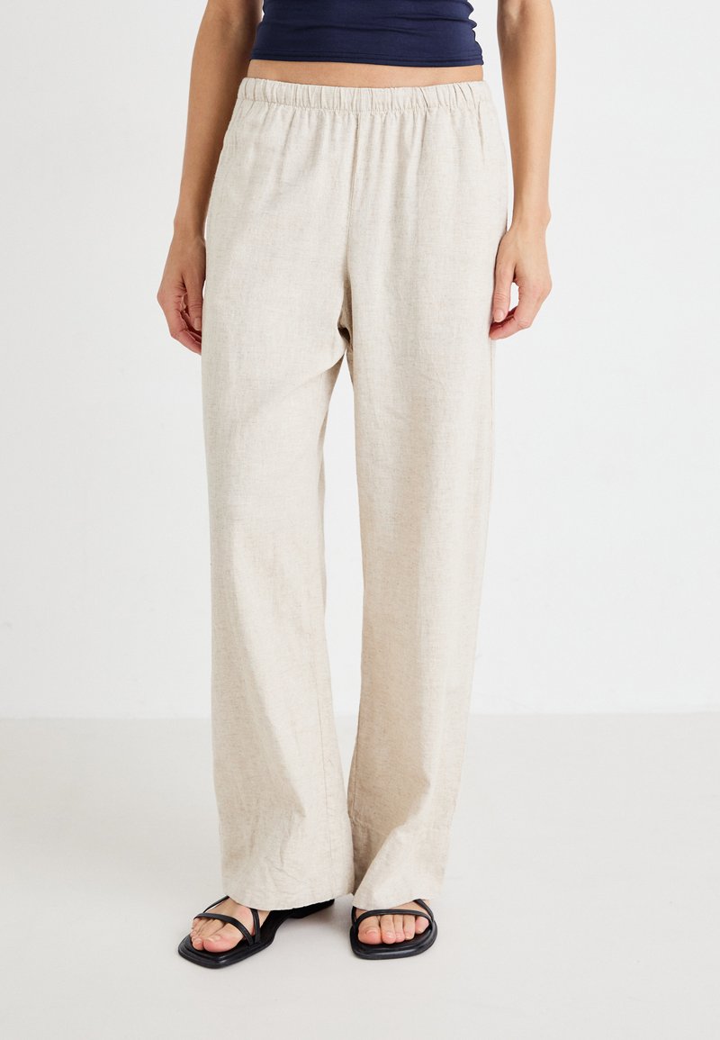 Pantalons en lin beige avec une taille élastique, des jambes larges, et une texture douce. Associés à des sandales plates noires.