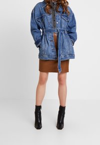 Jeansjacka med bälte i midjan, med stora fickor, avslappnad passform och blekt blå färg. Bärs över en brun läderkjol och svarta ankelboots.