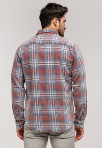 Mężczyzna odwrócony tyłem, ubrany w koszulę w kratę z długim rękawem w odcieniach czerwieni, niebieskiego i białego oraz jasnoszare spodnie, na jednolitym tle.