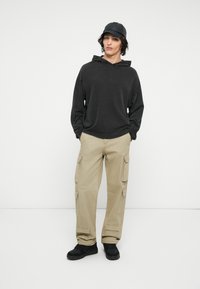 Sweat à capuche noir, pantalon cargo beige avec poches latérales, casquette noire et baskets noires. Tenue décontractée sur un fond uni.