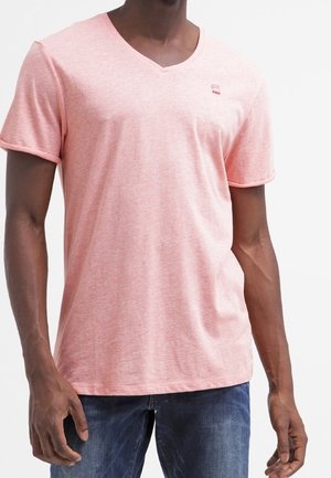 Mand iført en lyserød V-hals T-shirt med korte ærmer og blå jeans, står foran en hvid baggrund.