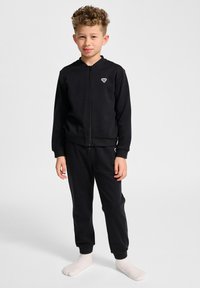 Zwarte zip-up hoodie en bijpassende broek van zachte stof, met elastische boorden en een klein logo op de borst. Model staat op blote voeten.