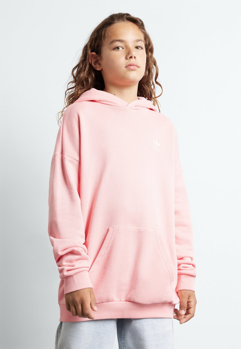 Sudadera con capucha rosa de corte relajado, que presenta un bolsillo frontal y una textura suave. Pequeño logo blanco en el pecho. Fondo liso.
