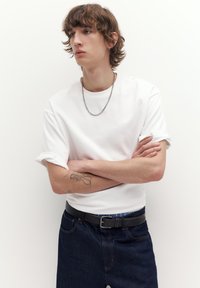 Junge Person mit lockigem Haar, die ein weißes T-Shirt mit hochgekrempelten Ärmeln, eine silberne Kettenkette, dunkle Jeans und einen schwarzen Gürtel trägt, die Arme verschränkt und ein Unterarmtattoo hat.