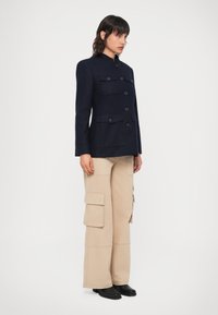 Tengerészkék gyapjú keverék dzseki magas gallérral, elöl gombos záródással és két mellzsebbel; bézs cargo nadrággal és fekete cipővel párosítva.