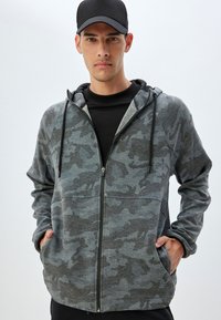 Sudadera con capucha de cremallera en camuflaje gris, con una textura suave, mangas largas, capucha con cordón y bolsillos laterales. Lleva puesta una camiseta negra debajo.