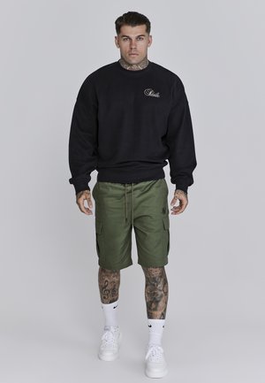 Schwarzer Pullover mit gerippten Bündchen, kombiniert mit olivgrünen Cargo-Shorts mit Seitentaschen. Weiße Sneakers mit sichtbarem Branding.