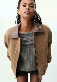 Brun hørflet jakke med en brun læderkrave, båret over en grå ribbet sweater, med lynlås og afslappet pasform.