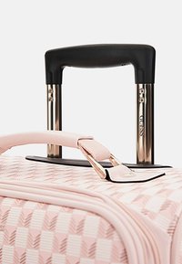 Valigia rigida rosa con motivo geometrico, manico telescopico nero e dettagli in hardware dorato. Superficie testurizzata e dettagli della cerniera visibili.