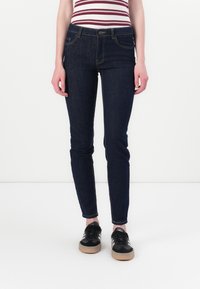 Άτομο που φοράει σκούρα μπλε στενά τζιν, ριγέ αμάνικο μπλουζάκι και μαύρα αθλητικά παπούτσια Adidas με λευκές ρίγες, στέκεται σε λευκό πάτωμα.