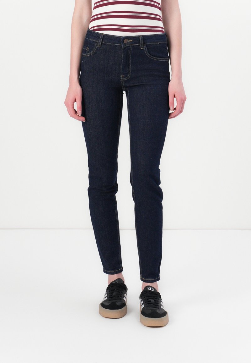 Άτομο που φοράει σκούρα μπλε στενά τζιν, ριγέ αμάνικο μπλουζάκι και μαύρα αθλητικά παπούτσια Adidas με λευκές ρίγες, στέκεται σε λευκό πάτωμα.