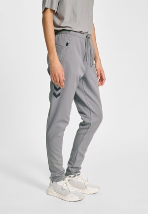 Person trägt hellgraue Jogginghosen mit Kordelzug am Bund und seitlichen Reißverschlusstaschen, kombiniert mit weißen Mesh-Sneakers vor einem schlichten Hintergrund.