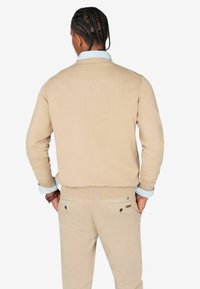 Beiger Pullover mit Rundhalsausschnitt, geripptem Saum und Bündchen, kombiniert mit beigen Hosen, die zwei Gesäßtaschen haben, und einem blauen gestreiften Hemd darunter.