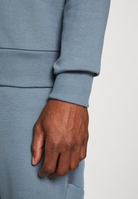 Polsino di felpa azzurro chiaro con texture a costine, caratterizzato da un design aderente. Una mano è appoggiata sul tessuto, mostrando il materiale morbido.