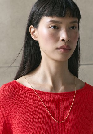 Young woman with long dark hair wears a bright red knit top and a thin gold necklace, looking slightly to the side against a neutral background.