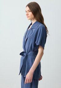 Blaues Leinen-Wickeljackett mit Kragen, kurzen Ärmeln und Taillengürtel. Glatte Textur, minimalistisches Design, in Seitenansicht gezeigt.