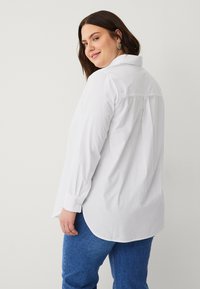 Camisa blanca de manga larga con cuello, puños abotonados y diseño con pliegue en la parte posterior, combinada con jeans azules, vista desde la parte de atrás.