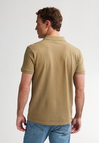Uomo con capelli castani corti, che indossa una polo beige a maniche corte e jeans blu, di spalle su uno sfondo chiaro e semplice.