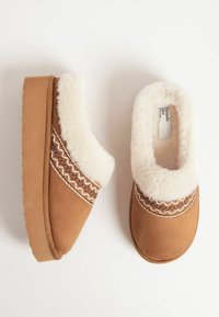 Barna suéd papucs fehér műszőrme béléssel. Díszes cikkcakk mintázattal rendelkezik a felsőrészén. Vastag talpa kényelmet és párnázottságot biztosít.