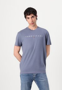 Kék pamut póló rövid ujjakkal, kerek nyakkivágással, fehér "TOMMY JEANS" logóval a mellkason, világoskék farmernadrággal párosítva.