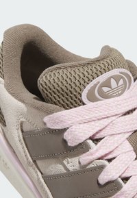 Brown suede sneaker featuring a mesh tongue, light pink laces, and a logo patch with three stripes design on the side. Textured accents visible.